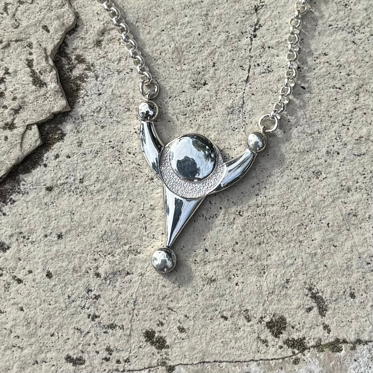 A silver pendant necklace with a stylized crop circle design, resting on a textured gray stone surface.