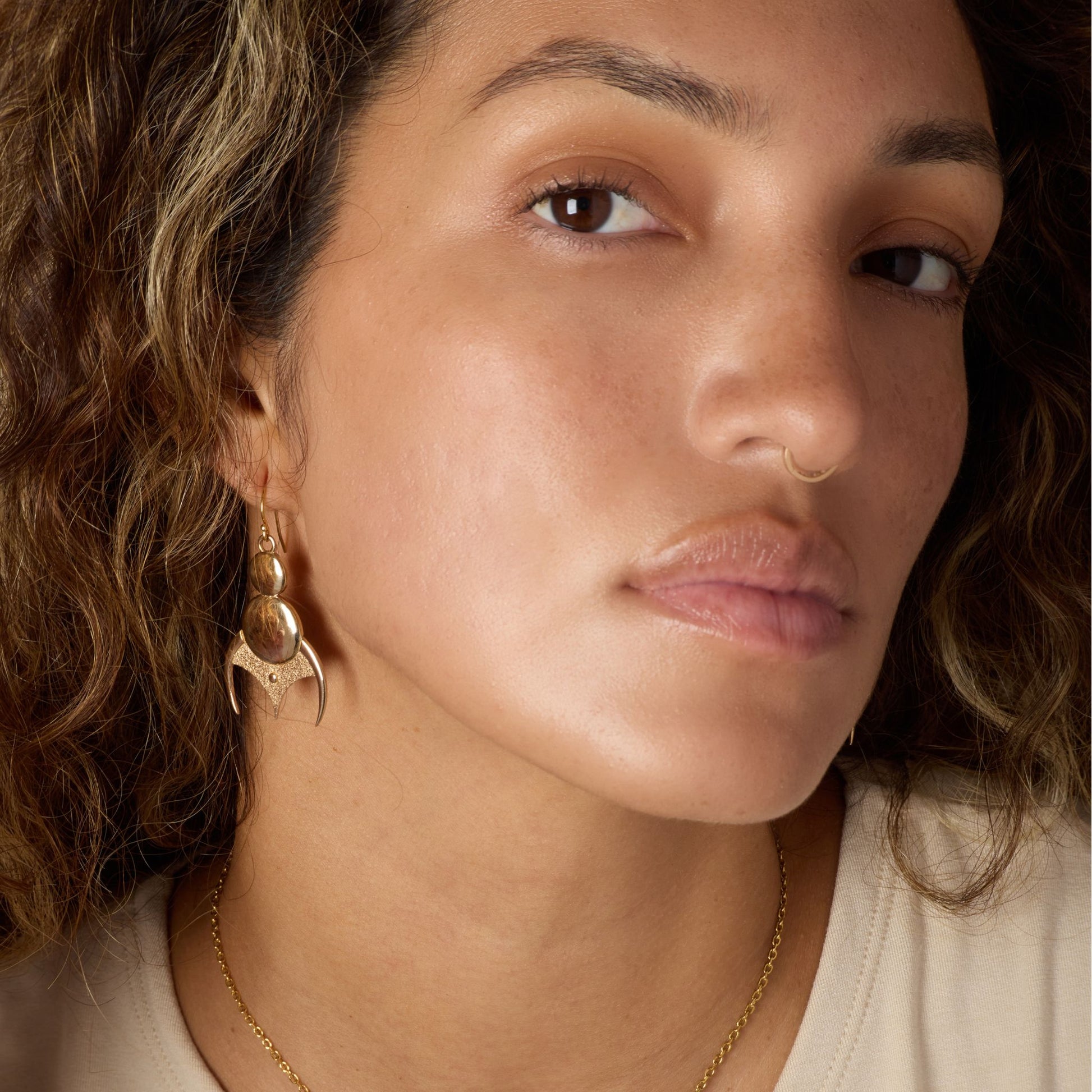 Close-up of a woman wearing gold-toned earrings and a nose ring, with a neutral background.