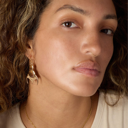 Close-up of a woman wearing gold-toned earrings and a nose ring, with a neutral background.