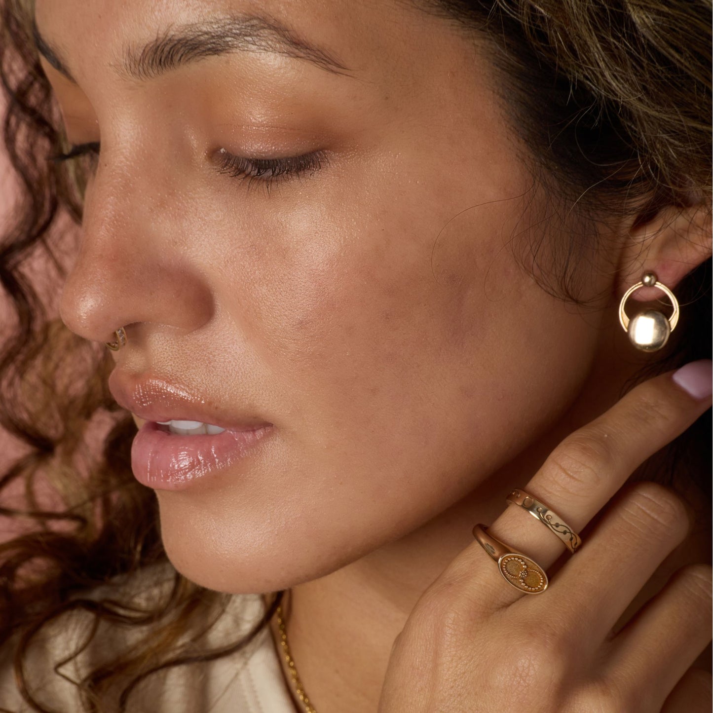 Close-up of a woman wearing gold-toned earrings and rings, with a soft focus on her face and hand.