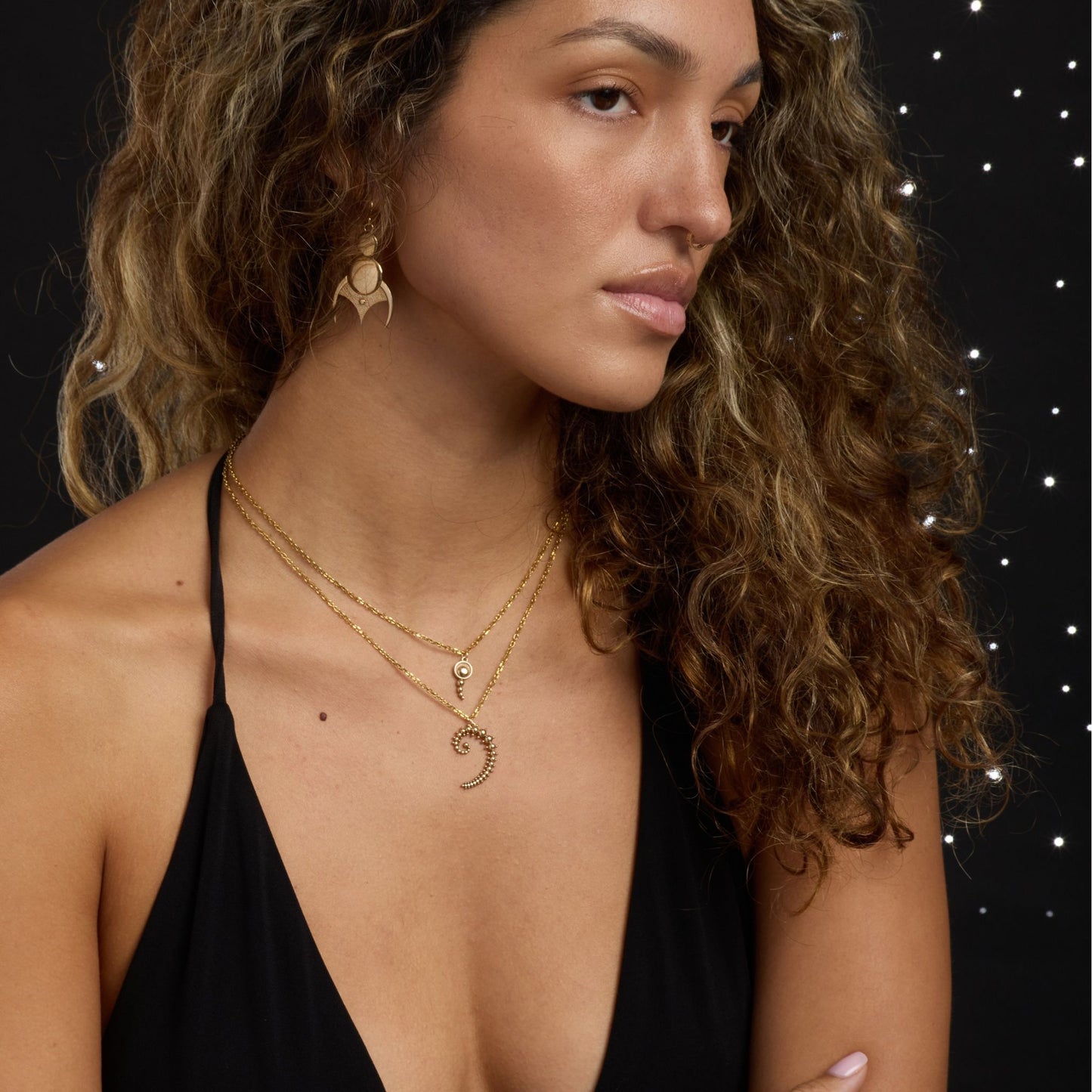 A woman with curly brown hair wearing layered gold-toned necklaces, including a fibonacci pendant, a small crop circle pendant, against a dark background with small white star-like specks.