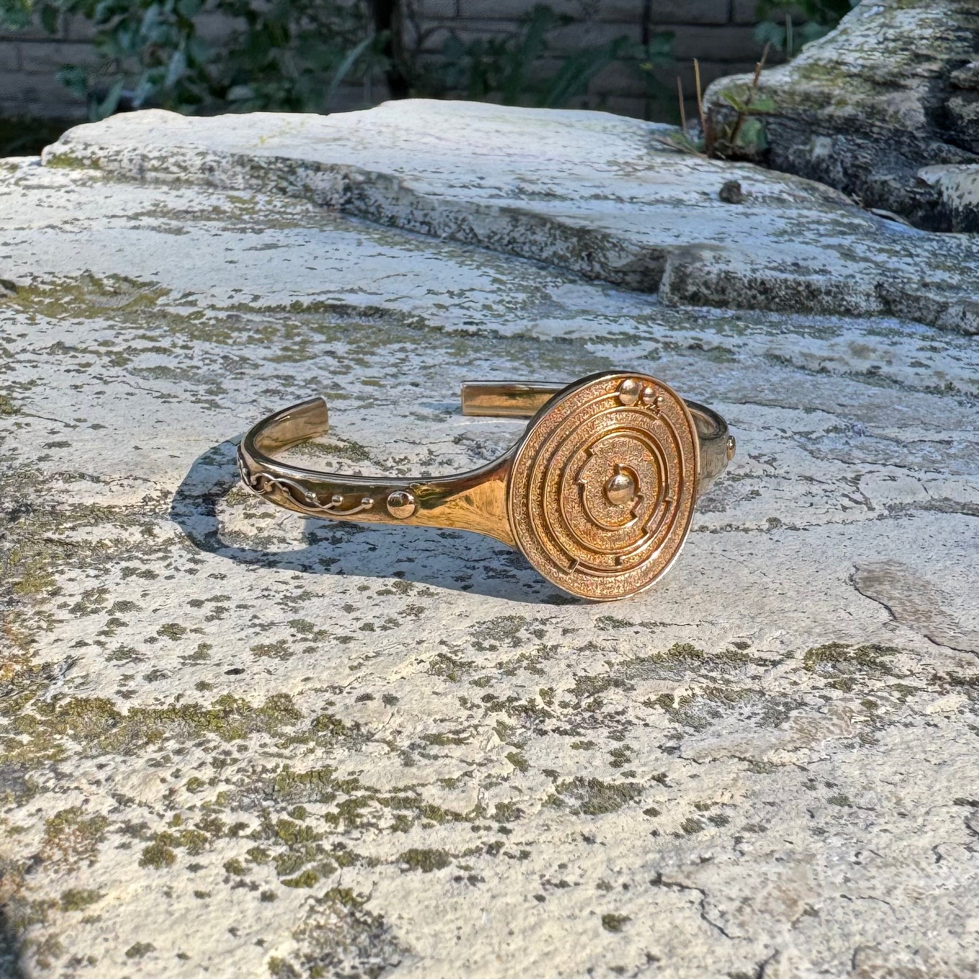 Gold-toned cuff bracelet with circular crop circle design on a stone surface