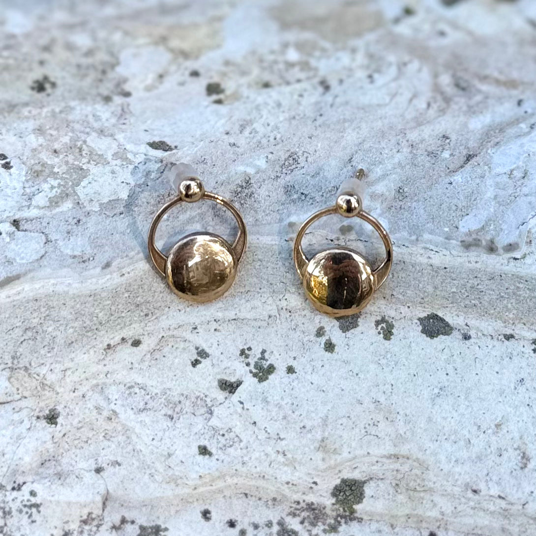 A pair of gold hoop earrings with a small polished disc at the center, displayed on a textured light gray stone surface.