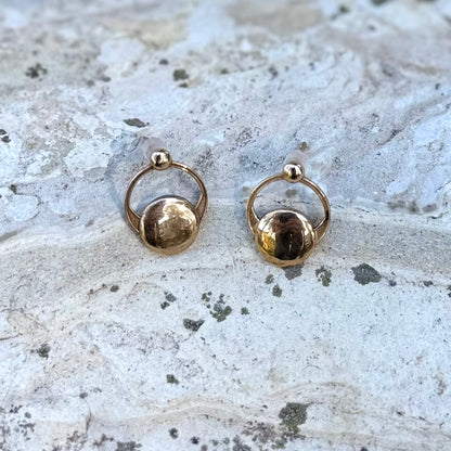A pair of gold hoop earrings with a small polished disc at the center, displayed on a textured light gray stone surface.