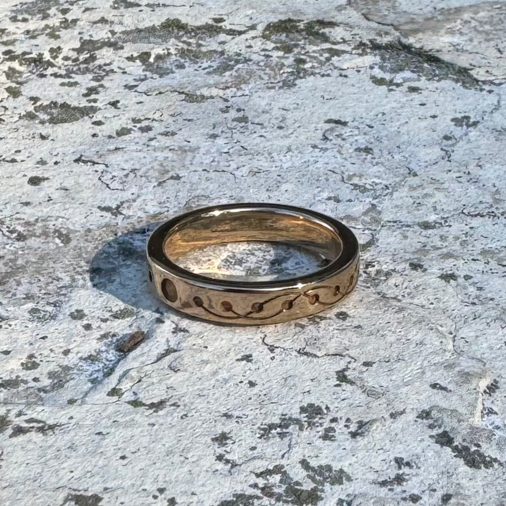 A gold-toned band ring with a wavy, crop circle design, resting on a textured, weathered stone surface.