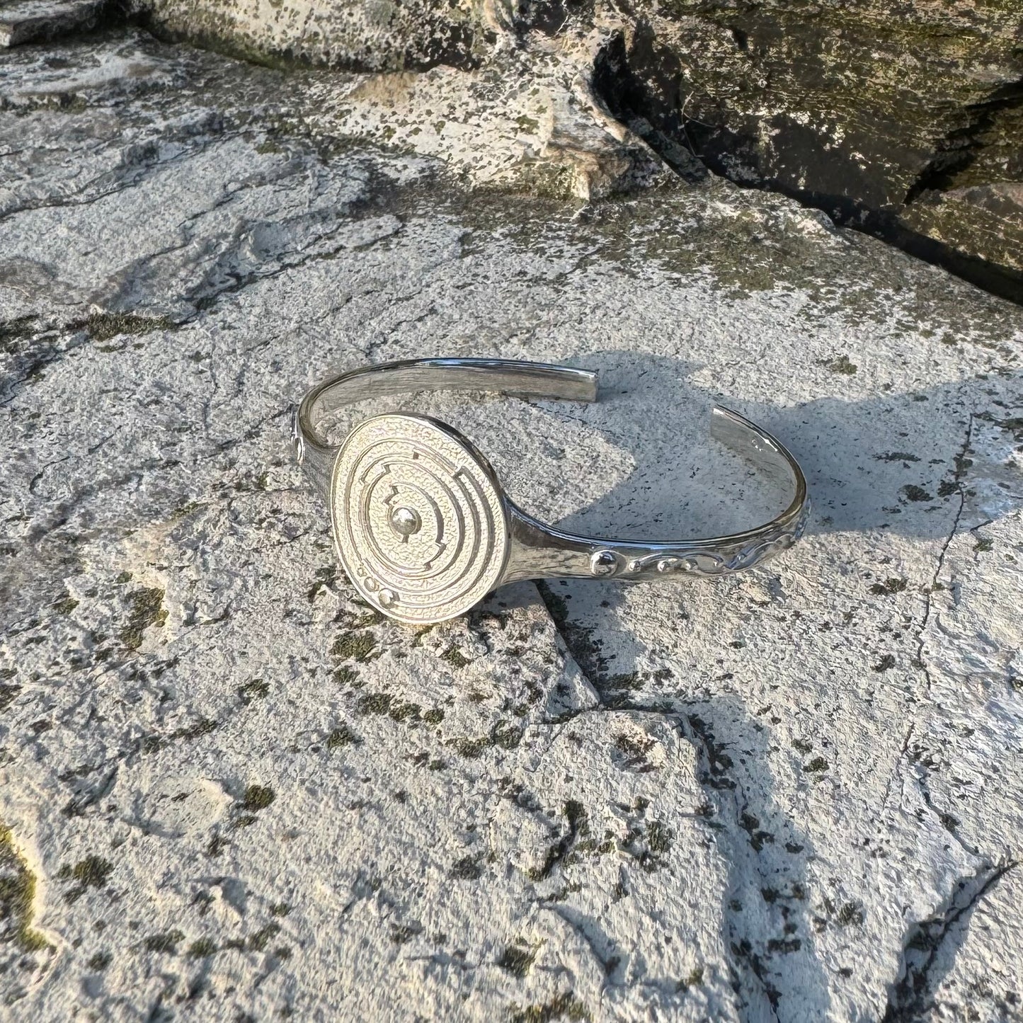 A polished silver cuff bracelet with a circular spiral design on one side, resting on a textured, mossy stone surface.