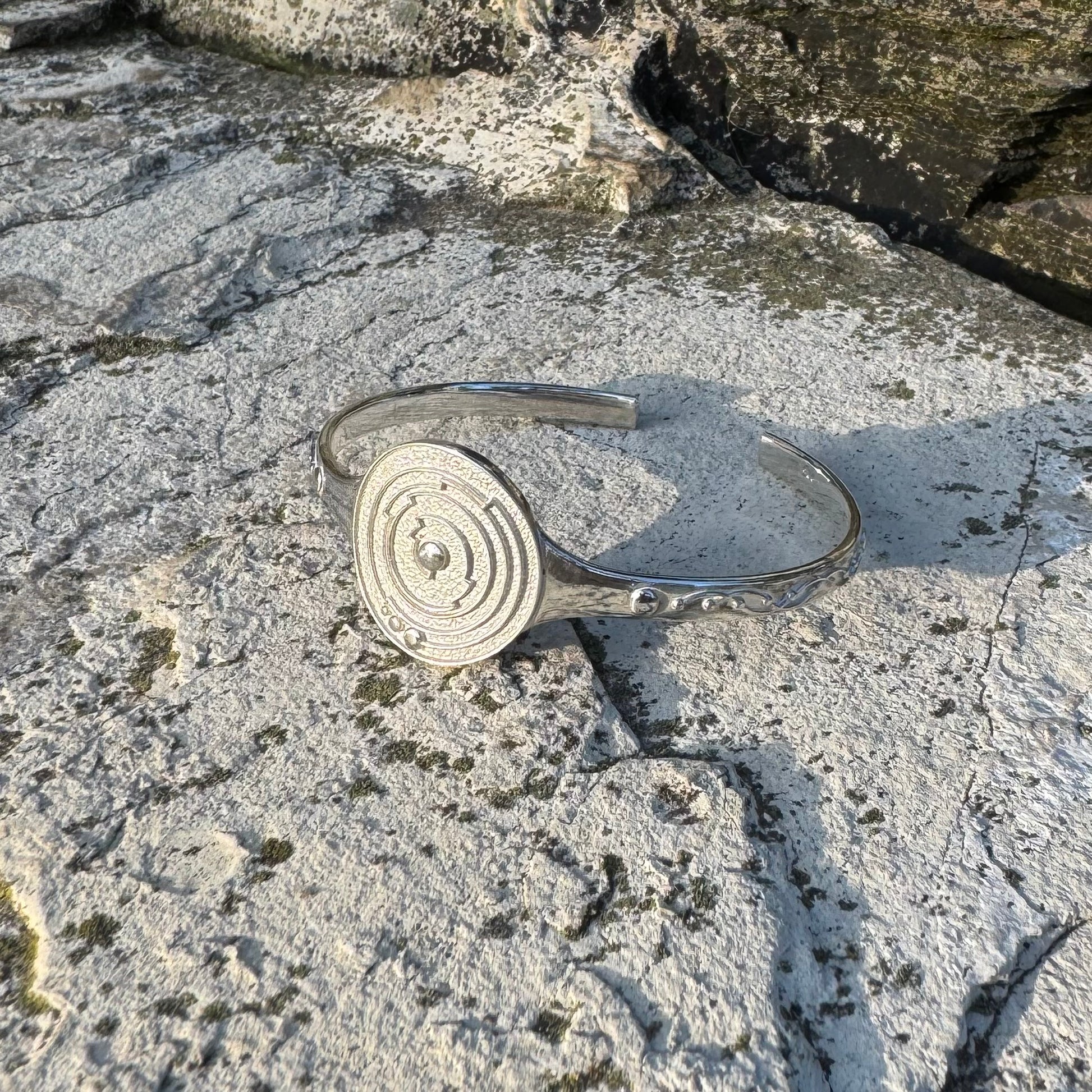 A polished silver cuff bracelet with a circular spiral design on one side, resting on a textured, mossy stone surface.