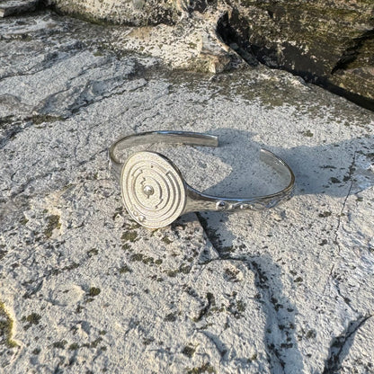 A polished silver cuff bracelet with a circular spiral design on one side, resting on a textured, mossy stone surface.