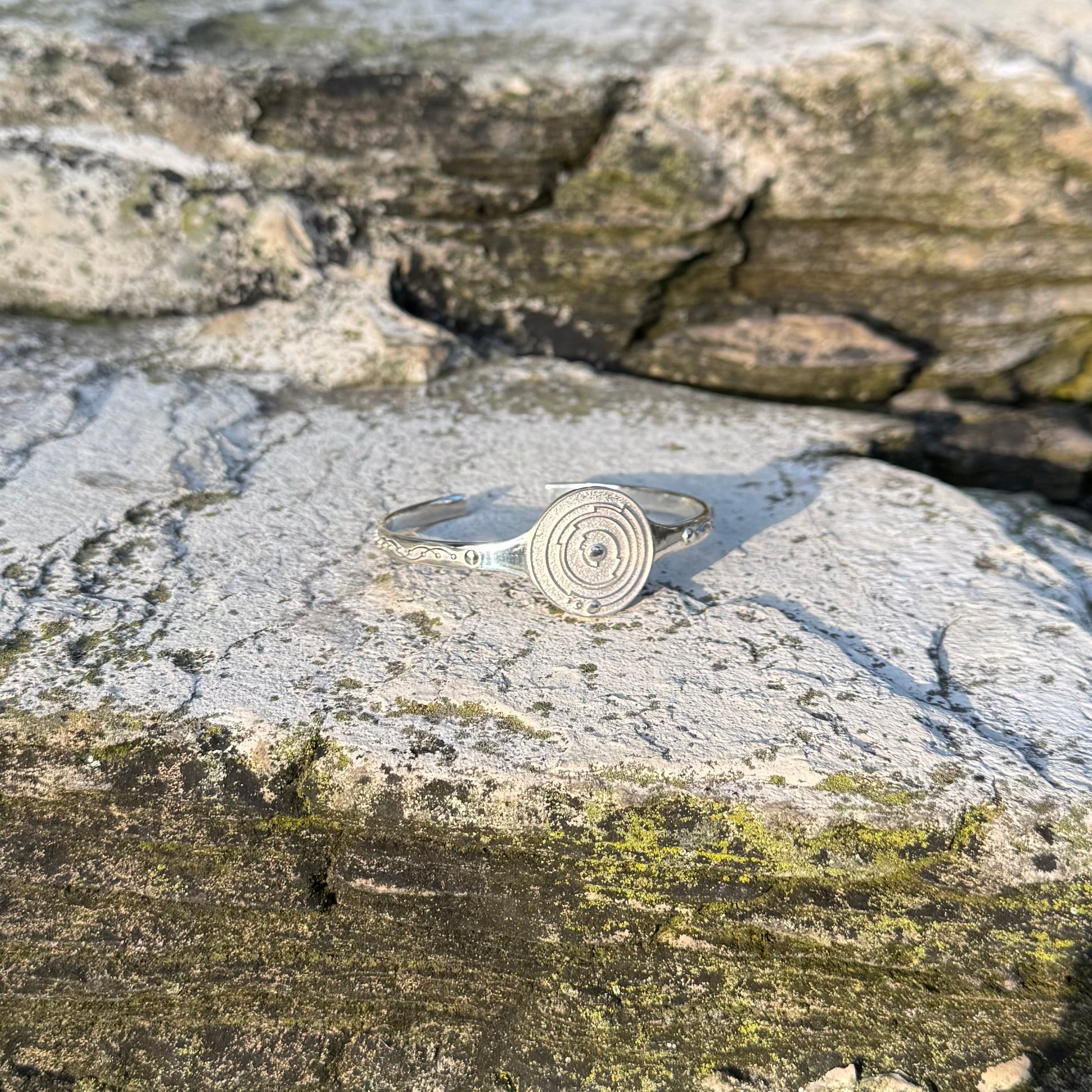 A silver cuff bracelet with a circular spiral design, resting on a textured, mossy gray stone surface.