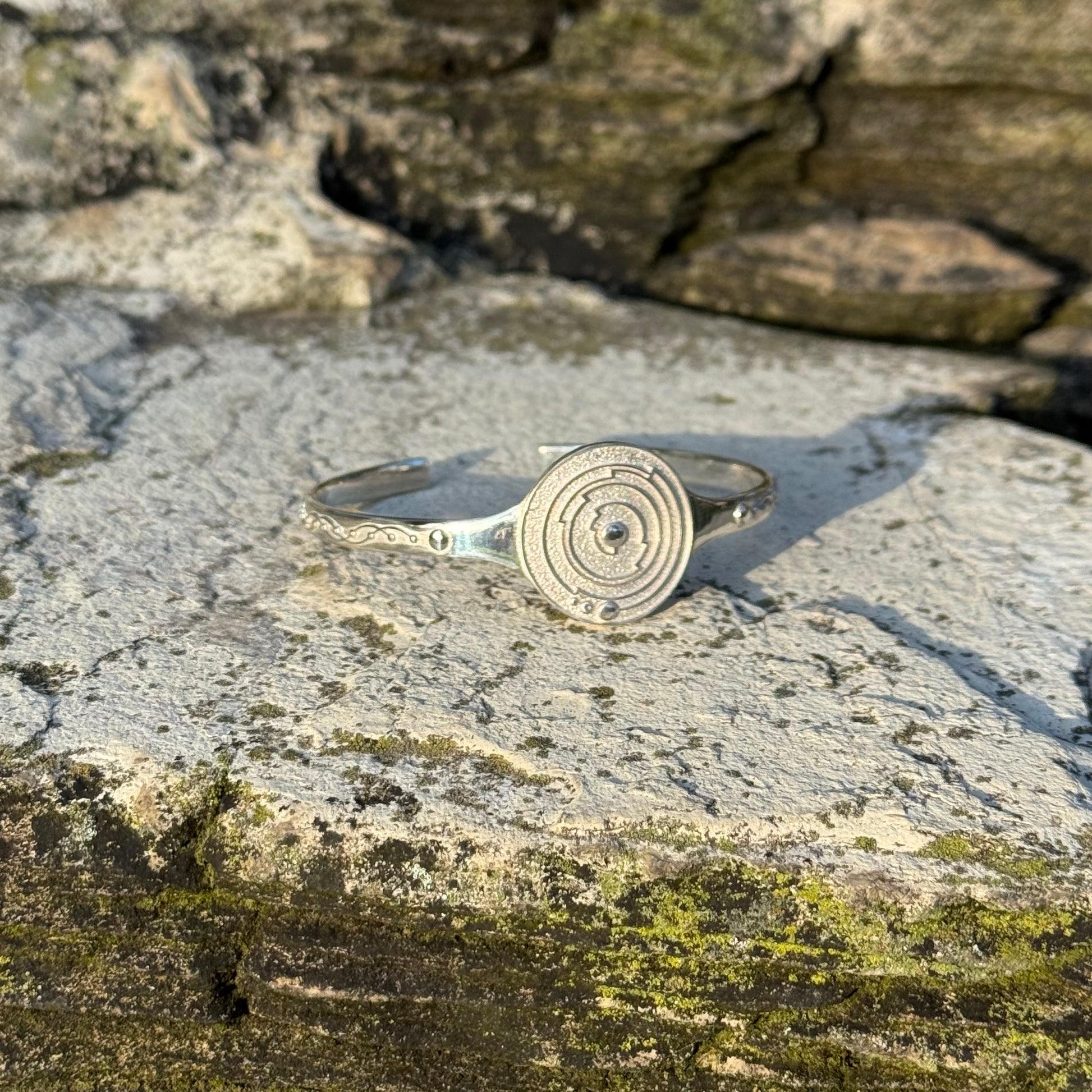 A sterling silver open cuff bracelet with a central spiral maze design, resting on a textured gray stone surface outdoors.