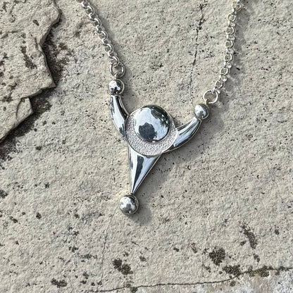 A silver pendant necklace with a stylized crop circle design, resting on a textured gray stone surface.