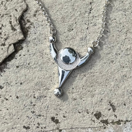 A silver pendant necklace with a stylized crop circle design, resting on a textured gray stone surface.