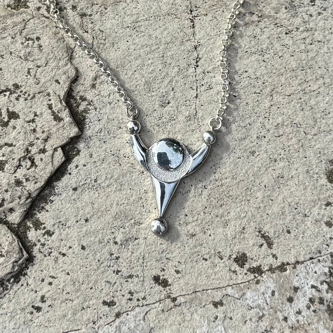 A silver pendant necklace with a stylized crop circle design, resting on a textured gray stone surface.