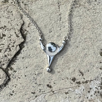 A silver pendant necklace with a stylized crop circle design, resting on a textured gray stone surface.
