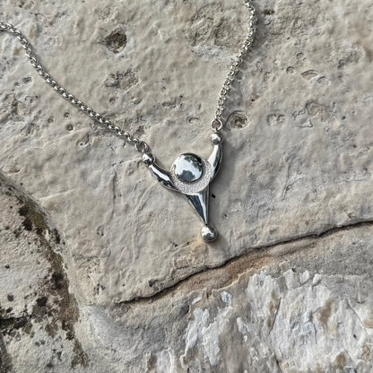 A silver necklace with a stylized pendant resembling a crop formation displayed on a textured light gray stone surface.