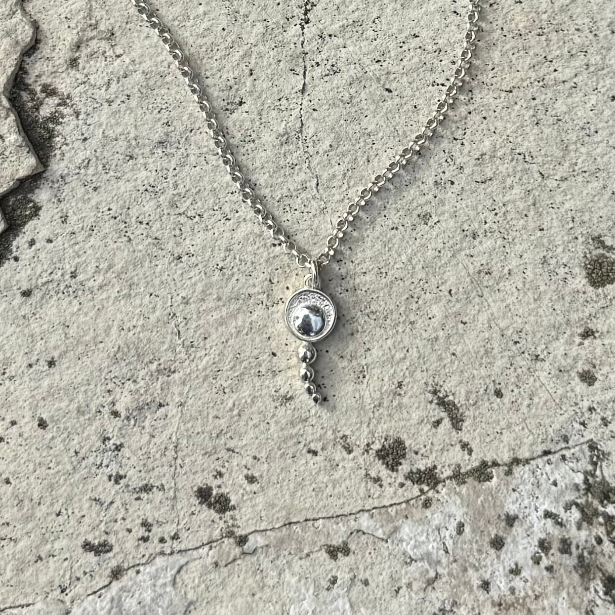 A silver chain necklace with a round pendant featuring a concentric circles, resting on a textured concrete surface.