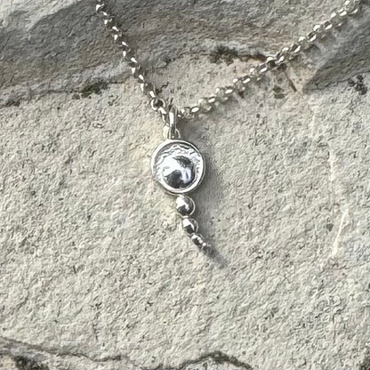 A silver chain necklace with a round pendant featuring concentric circles, with three dangling silver beads below, displayed on a textured gray stone surface.