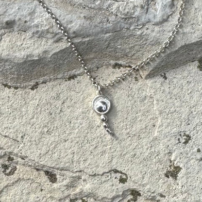 A silver chain necklace with a small circular pendant featuring a concentric circles, and a dangling bead, displayed on a textured gray stone surface.