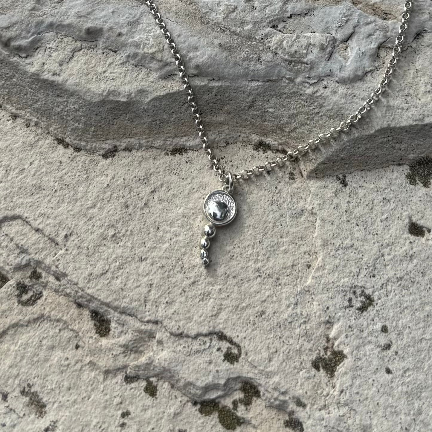 A silver necklace with small concentric circles pendant and dangling bead accents, laid on a textured gray stone surface.