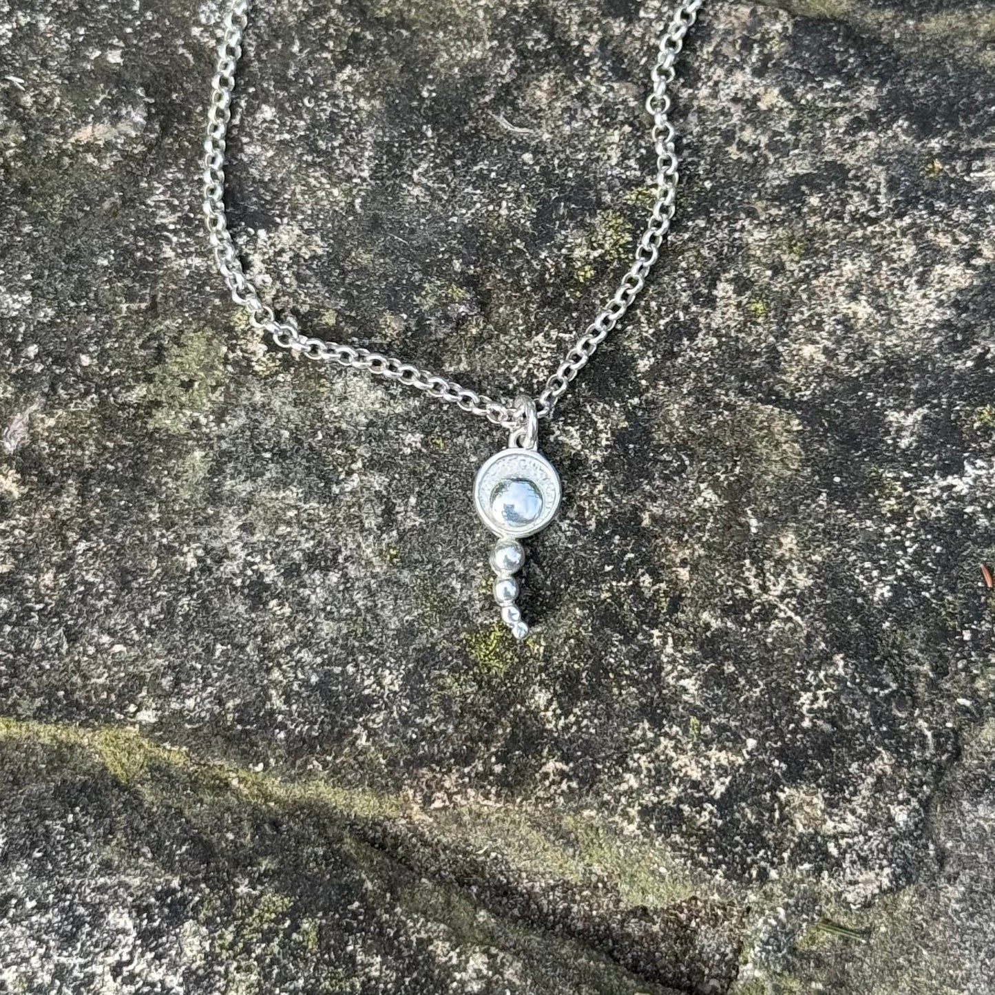 A silver-toned necklace with a circular pendant and small dangling beads, displayed on a textured stone surface.