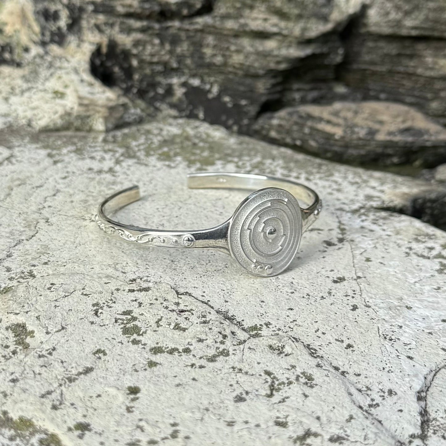 A silver cuff bracelet with a circular spiral crop circle design on the front and delicate leaf-like engravings on the band, resting on a textured gray stone surface.