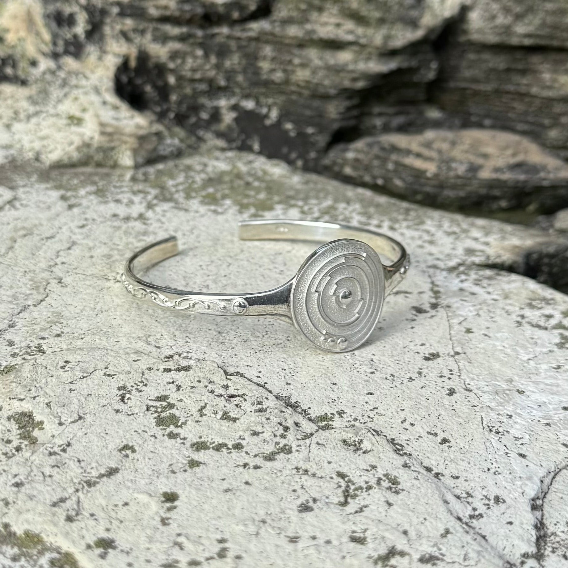 A silver cuff bracelet with a circular spiral crop circle design on the front and delicate leaf-like engravings on the band, resting on a textured gray stone surface.