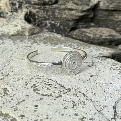 A silver cuff bracelet with a circular spiral crop circle design on the front and delicate leaf-like engravings on the band, resting on a textured gray stone surface.