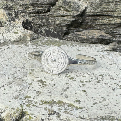 A silver metal cuff bracelet with a circular spiral crop circle design on the front, displayed on a textured gray stone surface with layered rock in the background.