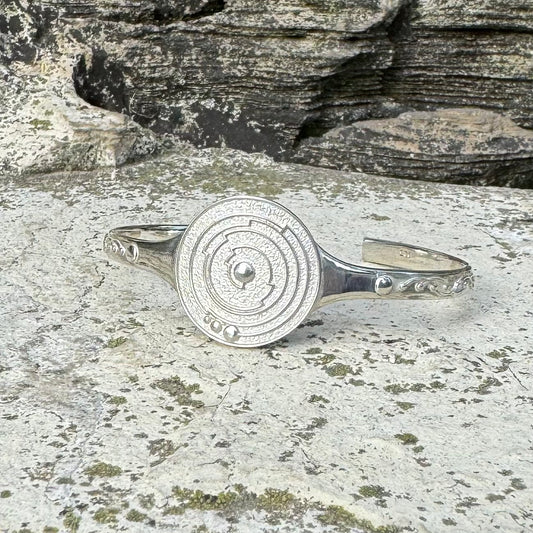 A polished silver cuff bracelet with a circular medallion featuring concentric crop circles and a central dot, resting on a textured stone surface with a rock face in the background.