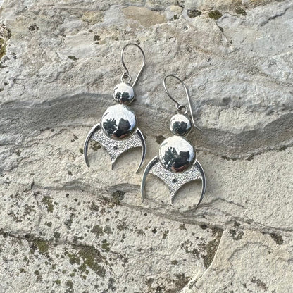 A pair of silver dangling earrings with polished and textured crescent moon and sphere details, displayed on a textured light gray stone surface.