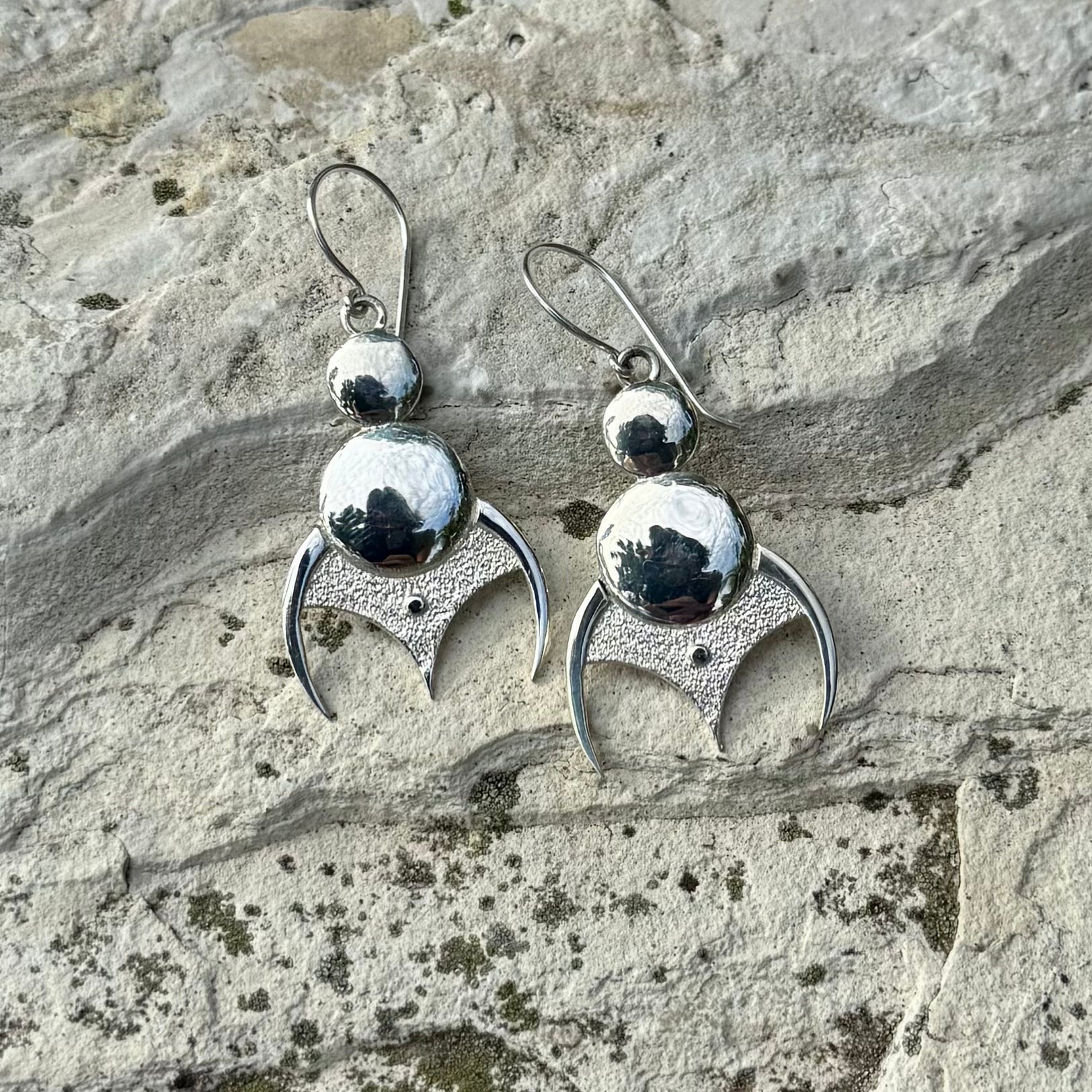 A pair of sterling silver dangle earrings with textured crescent moon shapes and polished round beads, displayed on a textured gray stone surface.