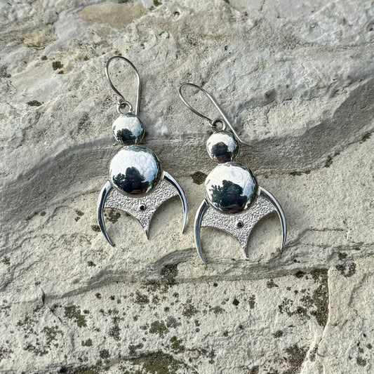 A pair of sterling silver dangle earrings with textured crescent moon shapes and polished round beads, displayed on a textured gray stone surface.