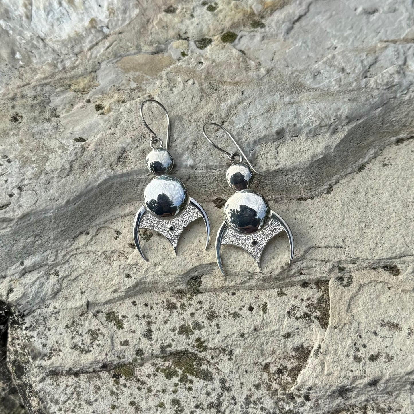 A pair of silver dangle earrings with polished and textured circular beads, resting on a textured gray stone surface.