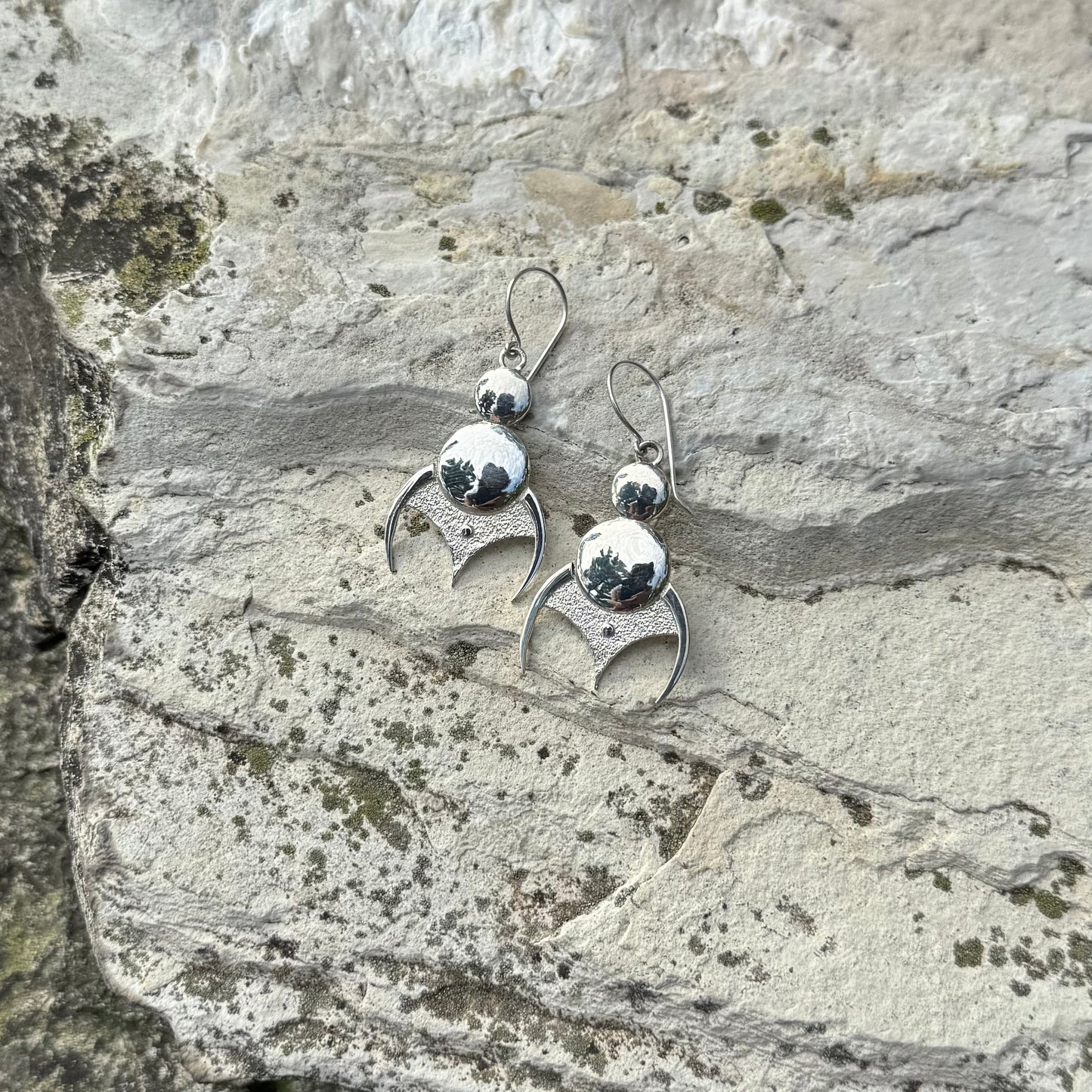 A pair of silver dangling earrings with a polished round bead and a textured crescent moon shape, displayed on a textured gray stone surface.