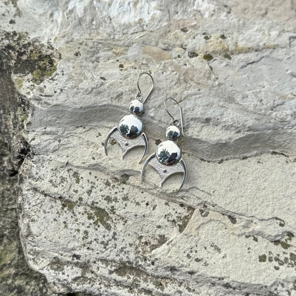 A pair of silver dangling earrings with a polished round bead and a textured crescent moon shape, displayed on a textured gray stone surface.