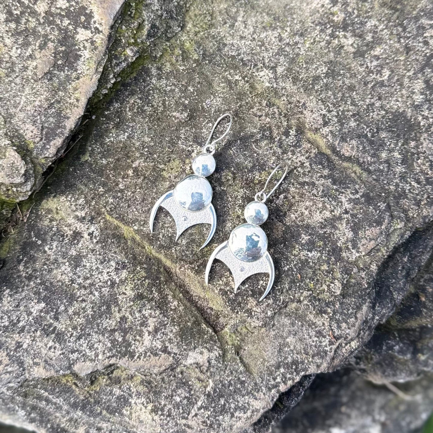 A pair of sterling silver dangle earrings with a curved, stylized pendant and a small round bead above, displayed on a textured gray rock surface.