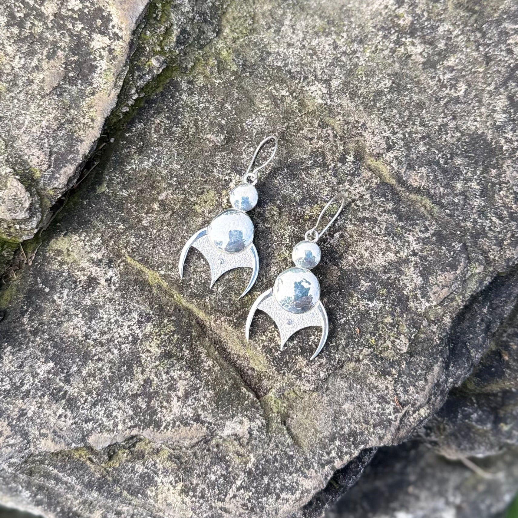 A pair of sterling silver dangle earrings with a curved, stylized pendant and a small round bead above, displayed on a textured gray rock surface.