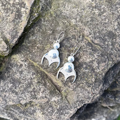 A pair of sterling silver dangle earrings with a curved, stylized pendant and a small round bead above, displayed on a textured gray rock surface.