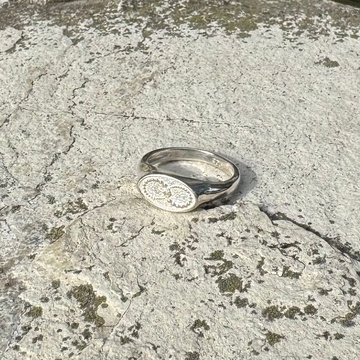 A silver signet ring with an oval face featuring an infinity crop circle, resting on a textured, mossy stone surface.