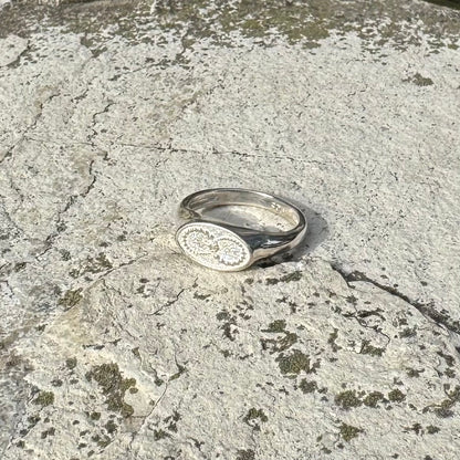A silver signet ring with an oval face featuring an infinity crop circle, resting on a textured, mossy stone surface.