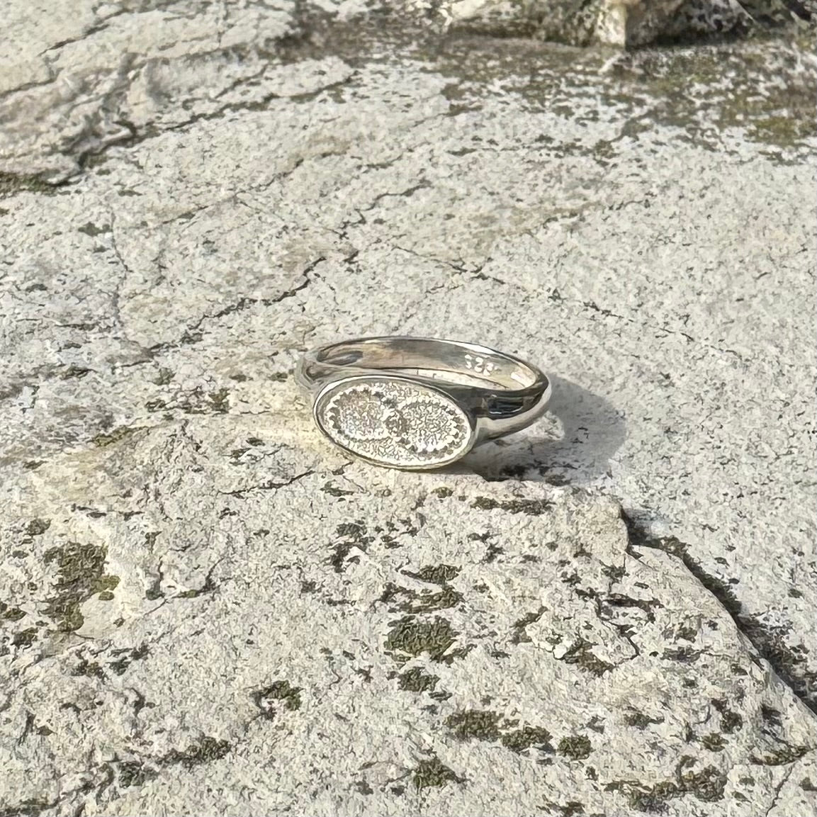 A silver signet ring with a textured oval face, featuring an infinity symbol, resting on a textured gray stone surface.
