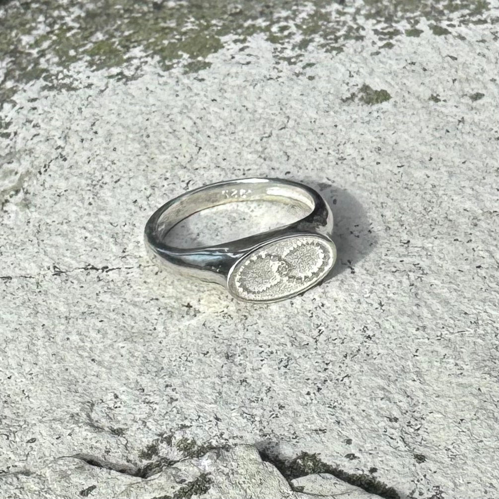 A silver signet ring with an oval face featuring a crop cirle motif with an infinity symbol, resting on a textured gray stone surface.