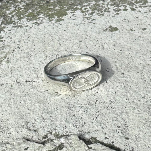 A silver signet ring with an oval face featuring a crop cirle motif with an infinity symbol, resting on a textured gray stone surface.