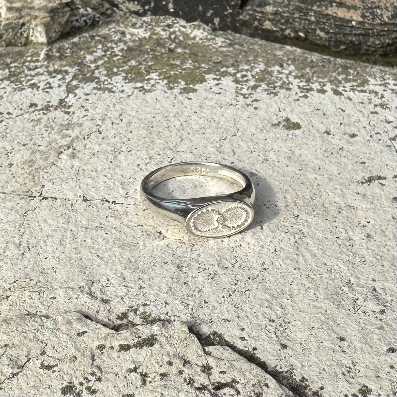 A polished silver signet ring with an oval face featuring a circular infinity symbol, resting on a textured, light gray stone surface.