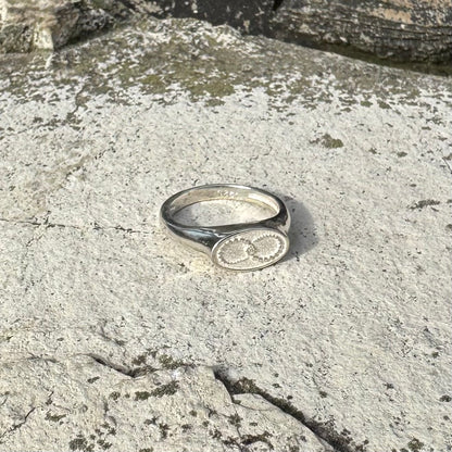 A polished silver signet ring with an oval face featuring a circular infinity symbol, resting on a textured, light gray stone surface.