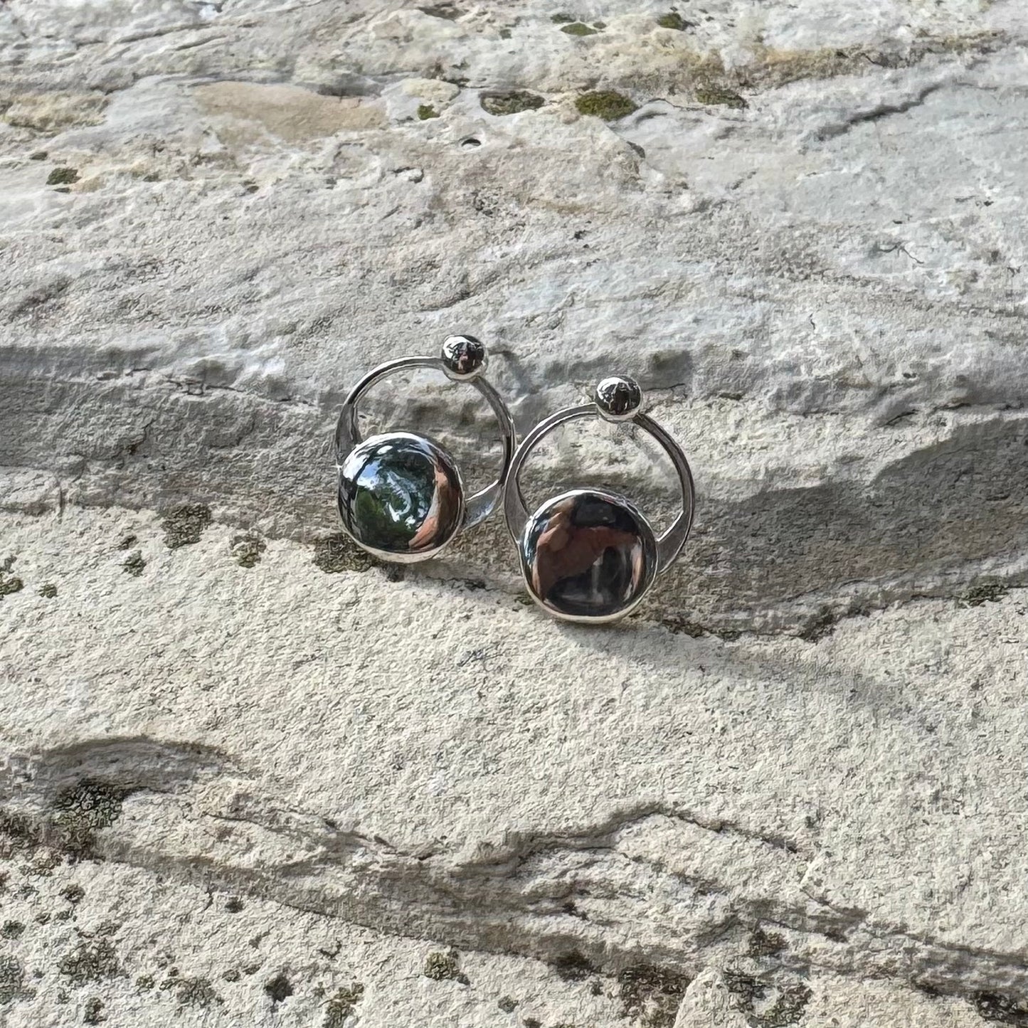 A pair of small silver hoop earrings with round circular discs, resting on a textured light gray stone surface.