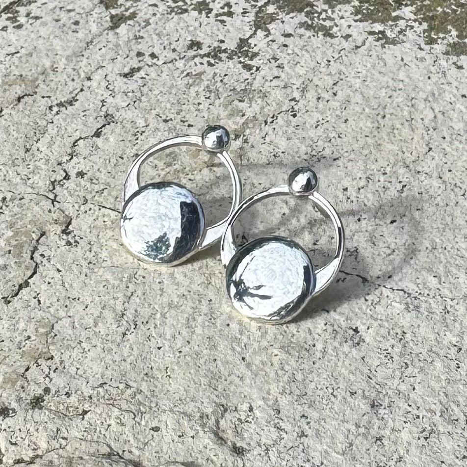 A pair of small polished silver hoop earrings with flat circular disc charms and small bead accents, photographed on a textured light gray stone surface.