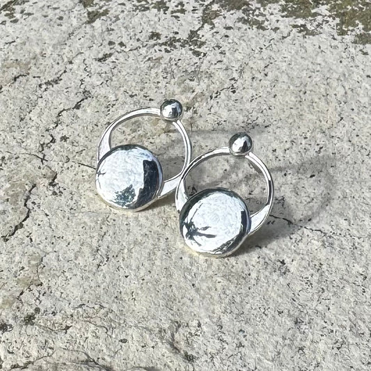 A pair of small polished silver hoop earrings with flat circular disc charms and small bead accents, photographed on a textured light gray stone surface.