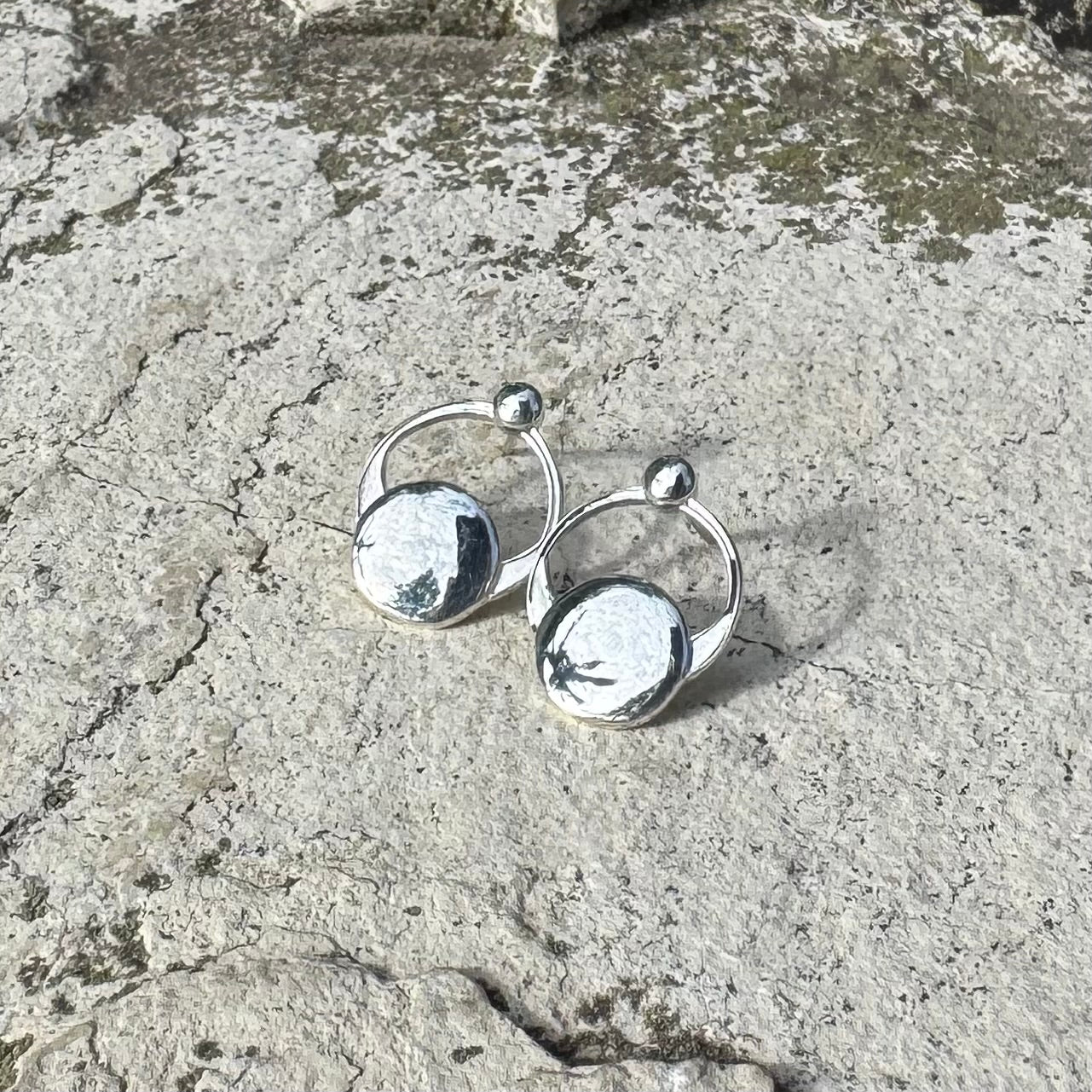 A pair of silver hoop earrings with circular disc charms, photographed on a textured concrete surface.
