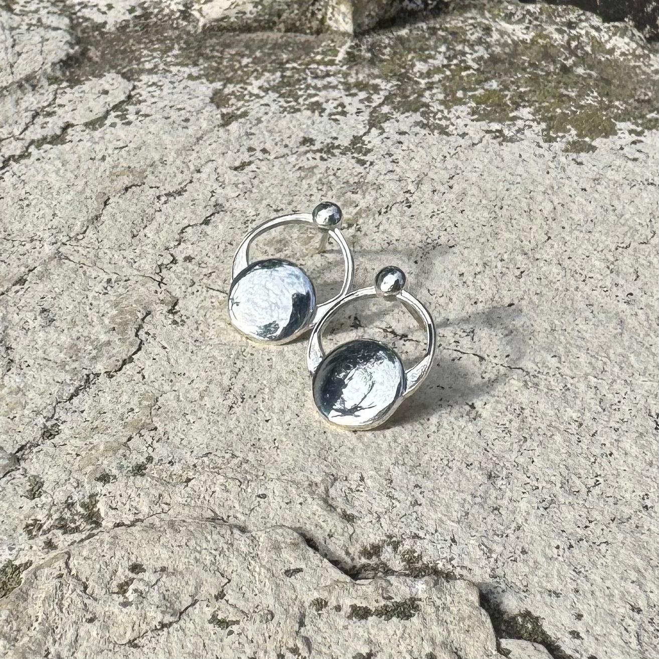 A pair of small silver  hoop earrings with flat circular disc charms and small bead details, photographed on a textured stone surface.